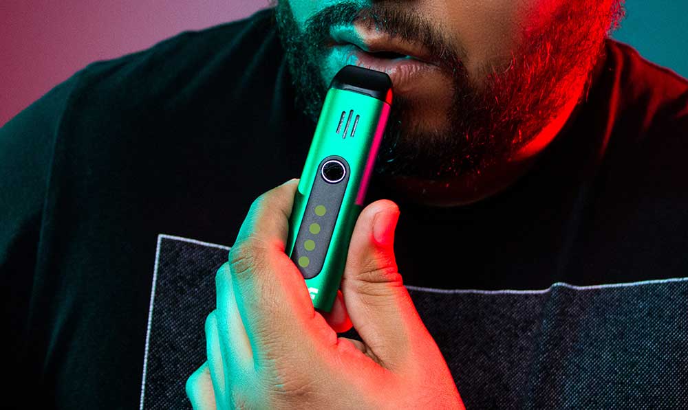 Person holding a teal WULF Flora portable dry herb vaporizer under red and teal lighting