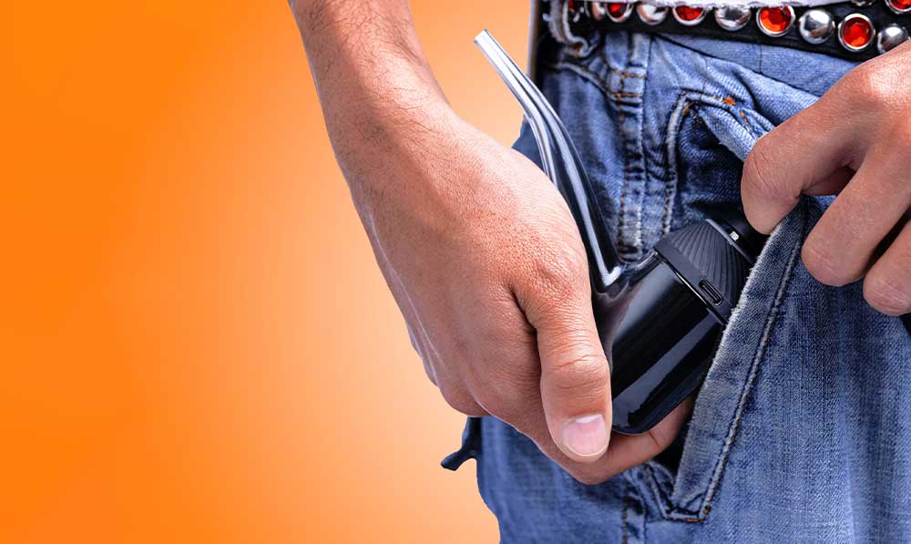 Hand slipping a Puffco Proxy vaporizer into a jeans pocket against an orange background