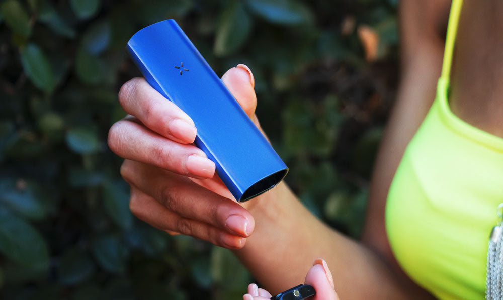 Woman holding periwinkle PAX Plus near bush trail