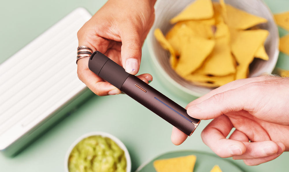 Two people passing Puffco Pivot Mocha over snack table with chips and guac