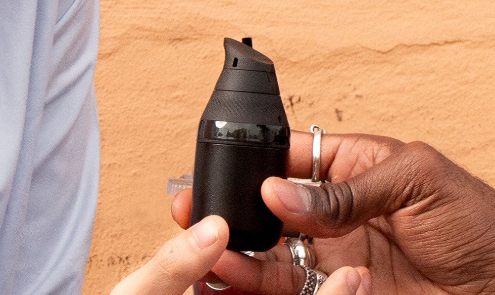 two hands sharing the PuffCo Proxy Core vaporizer in black on a orange/yellow background