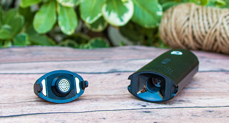 G pen Dash Vaporizer with mouthpiece opened on wooden table near bushes.