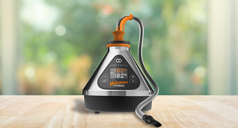 Volcano Hybrid Vaporizer with Tube attachment standing on wooden table with trees in background.