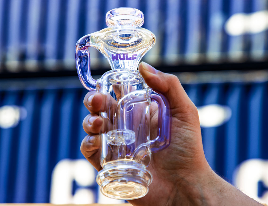 Hand holding a WULF purple and clear glass recycler bong in front of a blue shipping container